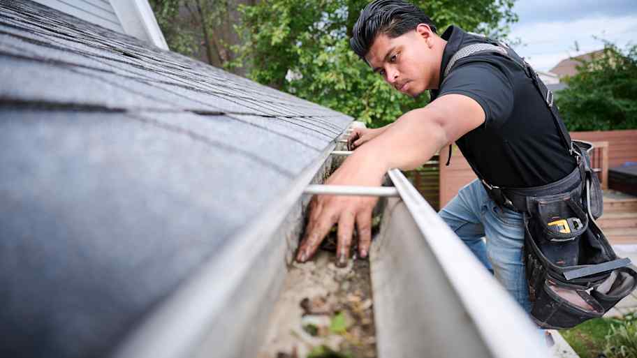Worker cleaning debris from a residential gutter by hand, wearing a tool belt and harness for roof safety maintenance.