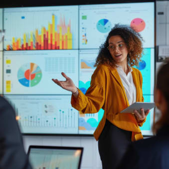 An African-American woman in a bright yellow blazer is standing in front of a screen displaying data. She gestures as she does a presentation, holding a tablet in her other hand. 