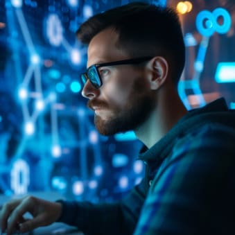 A bearded man with glasses is focusing on coding with two computer screens displaying blue and green lines of code against a blurred tech-themed background.