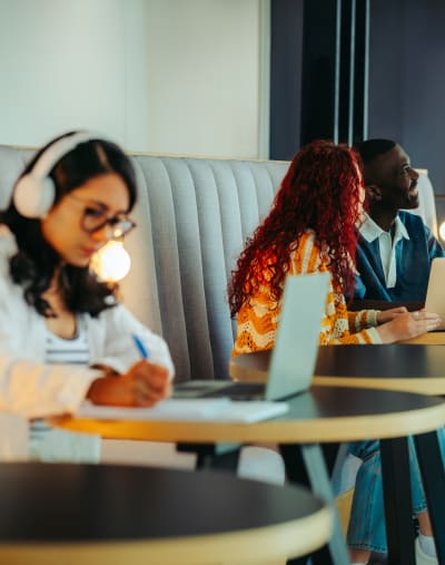 A diverse group of young adults in modern study lounge. They are holding study materials and using laptops; three of the students are engaged in conversation while another young woman works alone.
